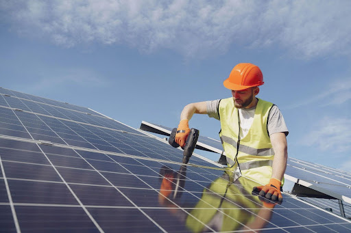 A man wearing an orange safety vest and a hard hat is installing a solar panel under clear blue skies.